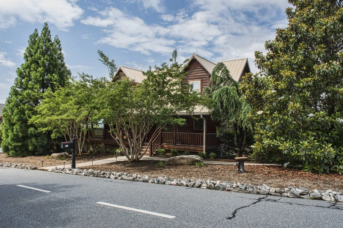 A wide-angle exterior view of the Bear's Den Cabin, featuring its log cabin facade, welcoming front porch, landscaped yard, and large trees in the background. The porch looks inviting for relaxing and the natural surroundings hint at nearby hiking and outdoor adventure.