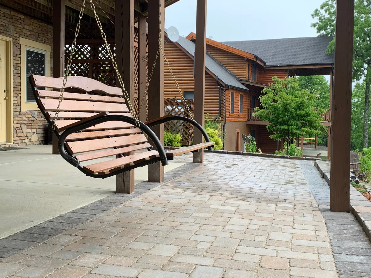 A swinging wooden bench positioned on the porch or patio area, with the cabin’s stone facade and wooden architectural elements visible, surrounded by landscaped greenery. The swing offers a tranquil spot to take in the sights and sounds of nature or chat with loved ones.