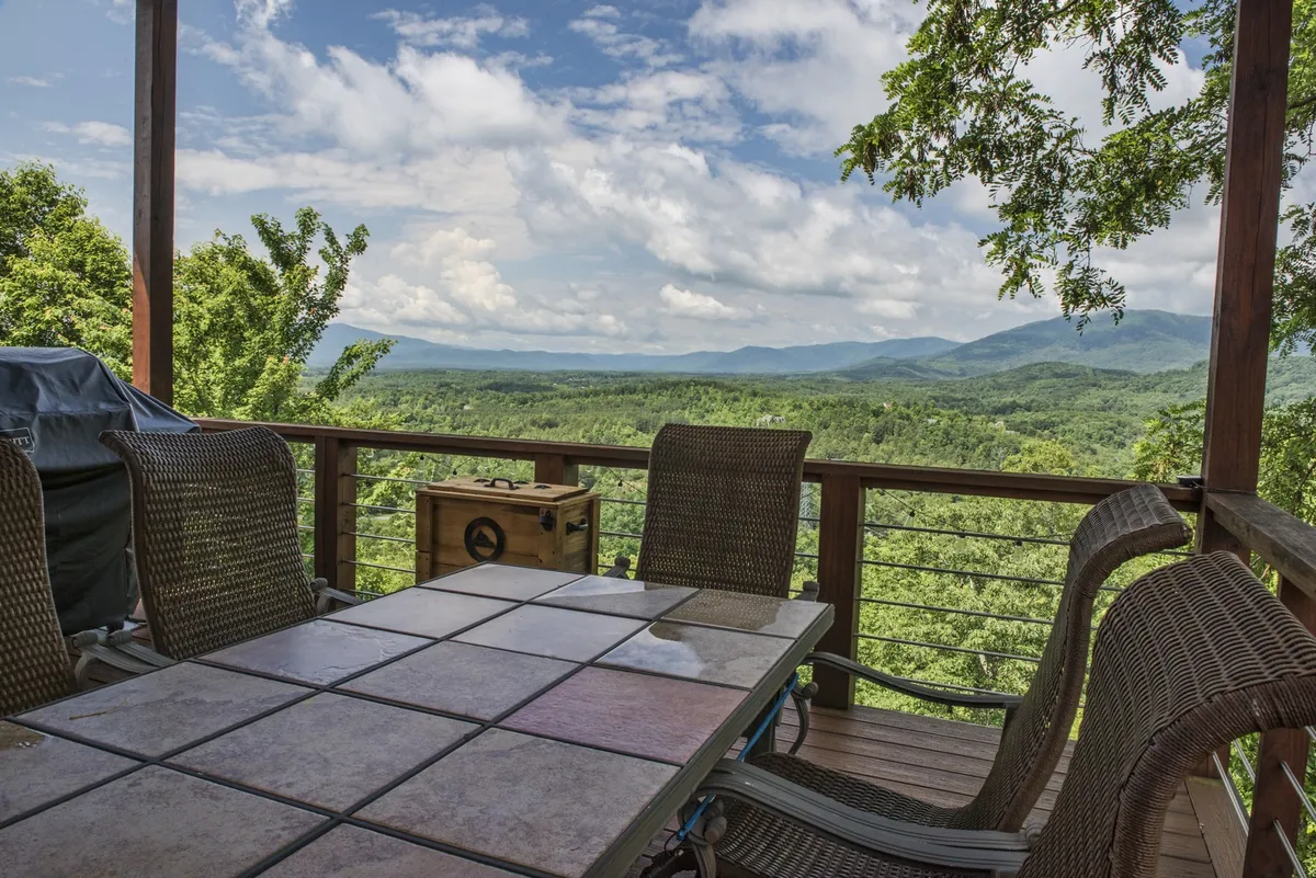 This image displays a spacious deck furnished with a dining table, outdoor furniture, and a grill, set against a stunning backdrop of Blue Ridge mountain views. The scene captures a tranquil, social atmosphere ideal for gatherings, outdoor dining, and relaxing while taking in the sunset.