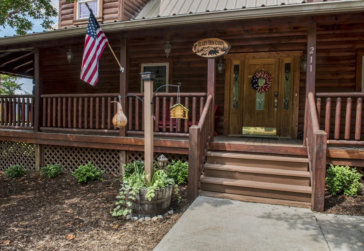 The image showcases the welcoming exterior of Bear's Den Cabin, featuring a charming wooden front door adorned with a 'welcome' sign, vibrant flower pots, an American flag fluttering by the entrance, and a cozy porch perfect for relaxing or watching sunsets.