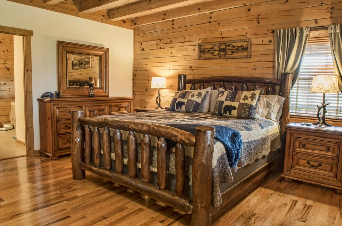 A warmly lit bedroom featuring a rustic log bed adorned with decorative pillows and soft blankets. A wooden dresser and nightstands flank the bed, complemented by nature-inspired decor that evokes a tranquil, mountain lodge feel.