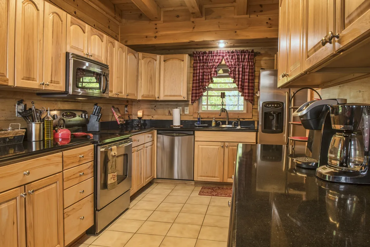 A view of the kitchen featuring wooden cabinetry, black countertops, stainless steel appliances, and a coffee maker by a window with plaid curtains. The space is well-equipped and welcoming, ideal for preparing meals or enjoying morning coffee rituals.