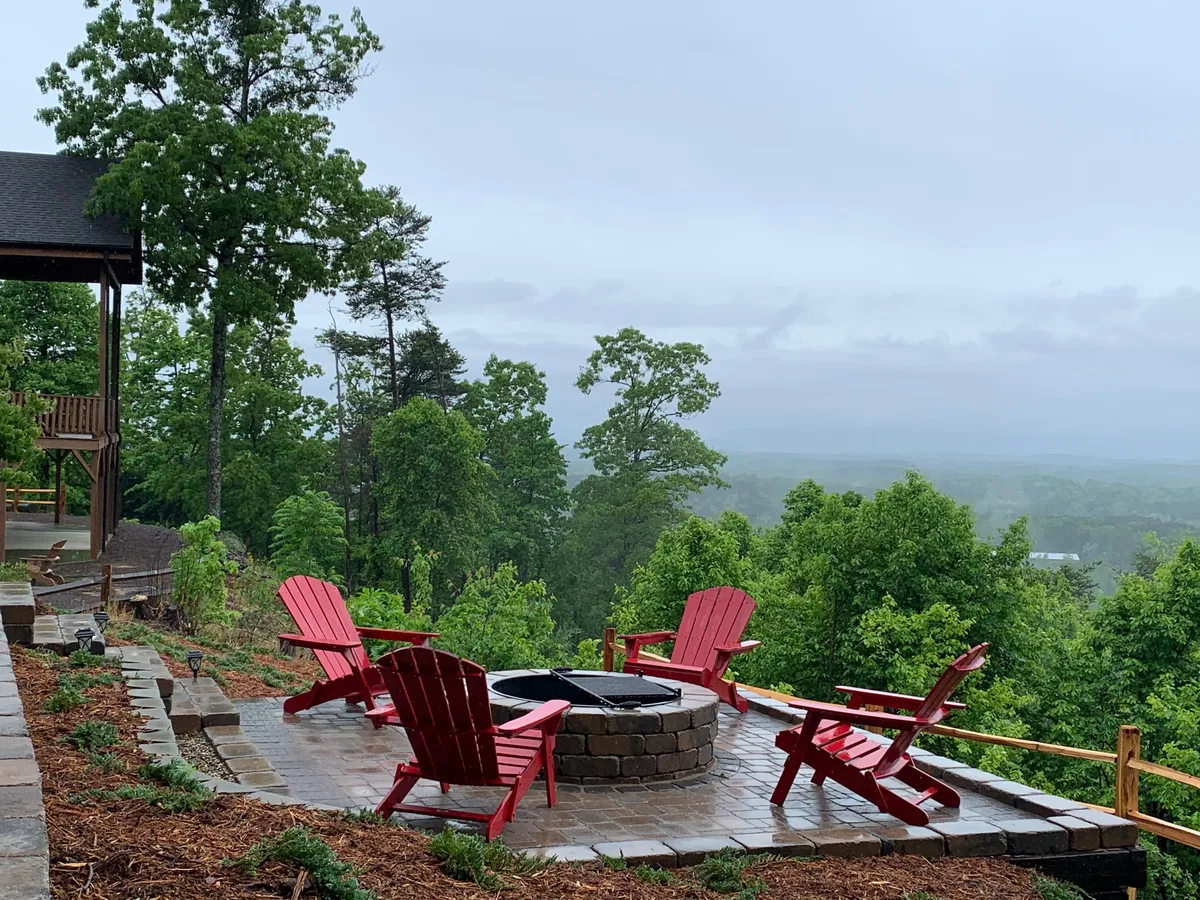 Two red Adirondack chairs encircle a stone firepit, surrounded by trees and distant hills. The cozy setup evokes evenings spent roasting marshmallows, sharing stories, and stargazing, with the natural landscape enhancing the serene, inviting mood.