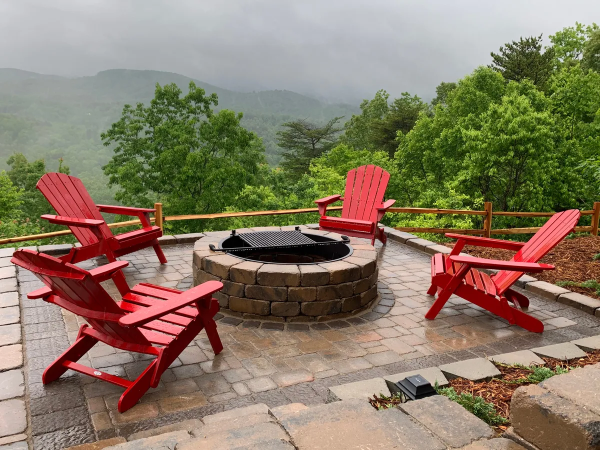 An outdoor fire pit area with red Adirondack chairs arranged in a circle, set amidst lush green surroundings and with a view toward rolling hills or mountains. The communal space is clearly designed for gathering, storytelling, and enjoying the outdoors together—especially in the evening.