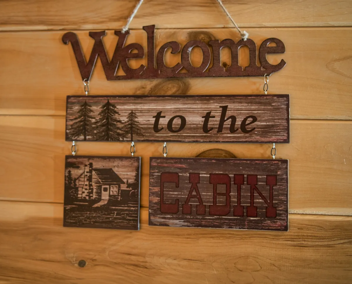 A welcoming sign against a wooden background, featuring an illustration of a cabin. The sign evokes an inviting, outdoorsy vibe and hints at the adventure and camaraderie to be found at Bear's Den Cabin.