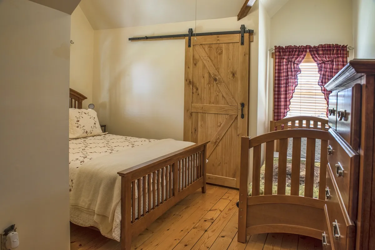 A spacious bedroom with a bed dressed in decorative bedding, a wooden barn door, and a baby crib positioned nearby. Checkerboard curtains and warm lighting create a family-friendly, inviting environment.